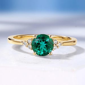 May include: A gold ring featuring a round, faceted green center stone held by four prongs. The band is accented on each side by a cluster of three small, clear, round-cut stones. The metal has a polished finish, and the design is symmetrical.