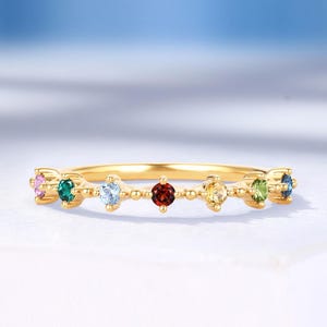 May include: A gold band ring featuring a series of colorful gemstones. The ring has a delicate design with small, round gemstones in various colors, including pink, green, blue, red, yellow, and green. The ring is set against a white surface.