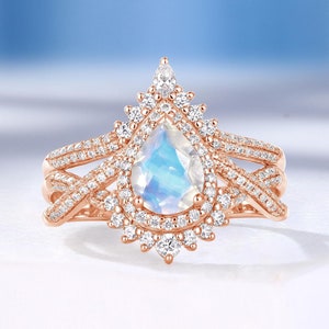 Peut inclure: Une bague de fian&ccedil;ailles en or rose avec une pierre de lune en forme de poire et un halo de petits diamants. La bague pr&eacute;sente un design d&eacute;licat et complexe avec une double bande.