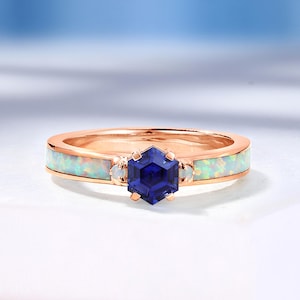 Puede incluir: Anillo de oro rosa con una piedra central hexagonal de color azul oscuro. La banda presenta incrustaciones de ópalo blanco. Dos piedras de ópalo blanco más pequeñas flanquean la piedra central. El anillo está sobre una superficie blanca con un fondo azul suave.
