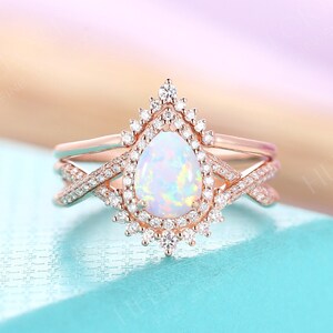 May include: A rose gold engagement ring with a pear-shaped opal gemstone surrounded by a halo of smaller diamonds. The ring is set on a delicate band with a criss-cross design.