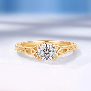 Könnte beinhalten: Ein goldener Verlobungsring mit einem runden, klaren Edelstein. Der Ring hat ein aufwendiges, graviertes Design am Band. Der Ring ist vor einem weißen Hintergrund mit einem sanften blauen Hintergrund platziert.