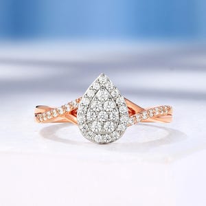 Può includere: Anello di fidanzamento in oro rosa con un gruppo di piccoli diamanti rotondi a forma di pera. La fascia ha un design attorcigliato con piccoli diamanti. L'anello è appoggiato su una superficie bianca con uno sfondo blu tenue.