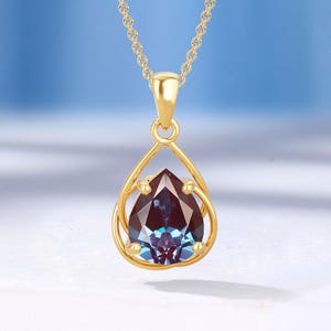 May include: A gold-colored pendant necklace featuring a teardrop-shaped gemstone with purple and teal hues. The gemstone is set within a delicate, open-frame gold setting, suspended from a fine gold chain. The necklace is displayed on a white surface.