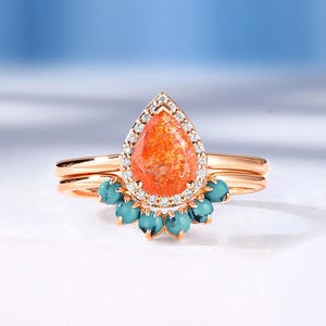 May include: A rose gold ring set featuring a pear-shaped orange sunstone center stone surrounded by a halo of small white diamonds. The band is accented with a curved row of five round turquoise cabochons set in a fan-like arrangement.