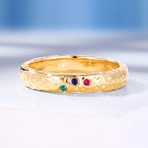 Puede incluir: Un anillo de banda de oro con una superficie texturizada y tres pequeñas piedras preciosas: verde, azul y roja. El anillo está sobre una superficie blanca con un fondo azul suave, mostrando joyería fina.
