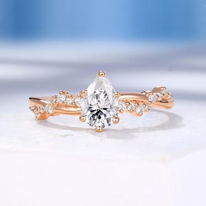 May include: A rose gold engagement ring with a pear-shaped diamond center stone. The band features a delicate, twisted design with small accent diamonds. The ring is set on a white surface with a soft blue background.