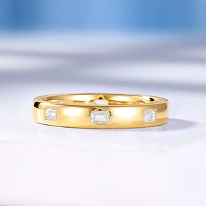 May include: A gold band ring featuring three evenly spaced, clear, rectangular-cut stones set flush into the metal surface. The ring has a polished, smooth finish and a consistent width, showcasing a clean, metallic yellow gold appearance throughout.