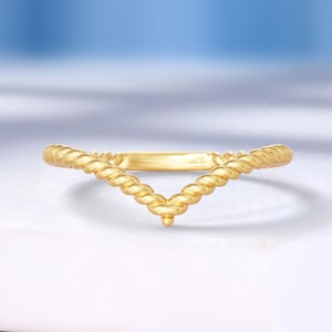 May include: A gold ring with a V-shaped design. The ring has a twisted rope-like texture along the band, with a small, pointed detail at the centre of the V. The ring is set against a white surface with a soft blue background.