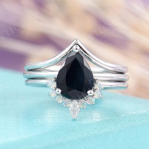 May include: A pear-shaped black sapphire engagement ring with a delicate, v-shaped band and a halo of smaller white gemstones. The ring is set on a blue background.
