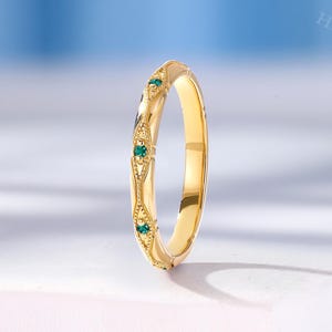 May include: A yellow gold band ring featuring a series of small, round, green gemstones set within decorative, diamond-shaped metal frames. The band has a polished finish and intricate milgrain detailing along the edges of the settings.