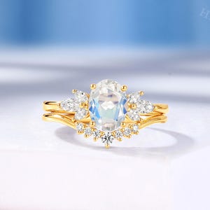May include: A gold ring set featuring an oval-cut moonstone center stone flanked by marquise-cut side stones. The band includes a curved nesting piece adorned with a row of small round-cut white stones, creating a layered, multi-stone design.