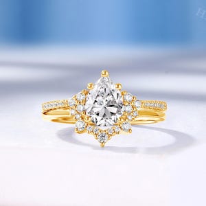 May include: A yellow gold ring featuring a central pear-shaped clear gemstone surrounded by a halo of smaller round clear stones. The band is thin and includes a row of pavé-set clear stones along the top half of the shank on both sides.