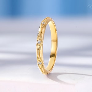 May include: A yellow gold band ring featuring a series of repeating diamond-shaped motifs along the outer surface. Each motif contains a small, round, clear gemstone set in the centre, accented by intricate milgrain detailing along the edges.