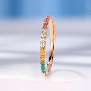 May include: A rose gold ring with a band of colorful gemstones. The gemstones are arranged in a rainbow pattern, transitioning from red to orange, yellow, green, and blue. The ring is set against a white surface with a soft blue background.