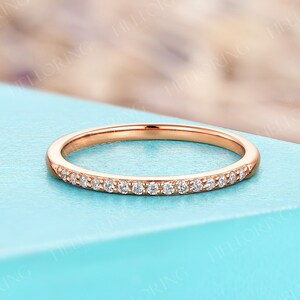 May include: Rose gold band ring with a row of small, clear gemstones. The ring is set against a light blue background. The ring is a delicate piece of jewelry.