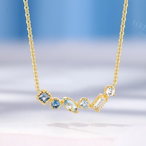 Op de afbeelding: Een gouden ketting met een cluster van zes blauwe en heldere edelstenen in verschillende vormen. De stenen omvatten een vierkante, twee ronde, een marquise, een rechthoekige baguette en een kleine ronde slijpvorm, gezet in goudkleurig metaal.