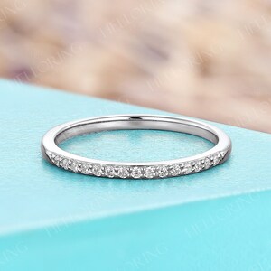 May include: A delicate silver ring with a band of small, clear gemstones. The ring is set against a light blue surface, with a blurred background of purple and white. The ring is a fine jewelry item.