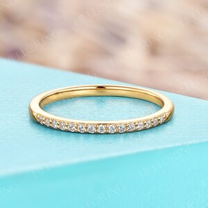May include: A delicate gold band ring with small, clear gemstones set along the top. The ring is displayed on a light blue surface. The HELLORING FINE JEWELRY logo is visible in the background.