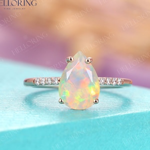 Puede incluir: Un anillo de compromiso de opal de forma de pera engastado en oro rosa con una banda de pequeños diamantes. El anillo está sobre una superficie azul claro.