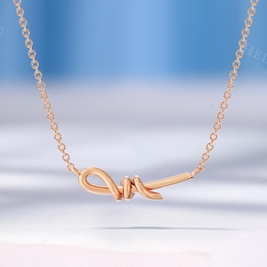 May include: A delicate rose gold necklace featuring a stylized knot pendant. The pendant is the focal point, suspended from a fine chain. The necklace is displayed on a white surface against a soft blue backdrop, highlighting the jewelry's elegant design.