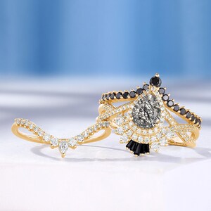 May include: A gold ring set with a pear-shaped clear gemstone surrounded by smaller clear gemstones and black gemstones. The ring is set on a white surface.