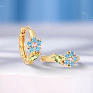 May include: A pair of gold hoop earrings. Each earring features a flower design with light blue and pink gemstones, along with a green leaf-shaped accent. The earrings are displayed on a white surface against a light blue background.