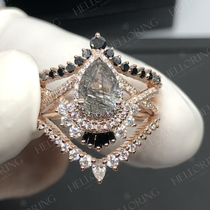 May include: A pear-shaped engagement ring with a grey and black gemstone surrounded by a halo of smaller diamonds. The ring is set in rose gold and features a delicate band with black diamonds.