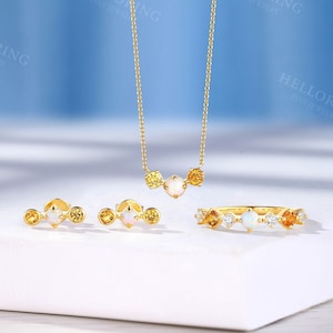 Puede incluir: Un conjunto de joyas de oro que incluye un collar, pendientes y un anillo. Las joyas están adornadas con pequeñas piedras preciosas redondas de color amarillo y ópalos blancos.