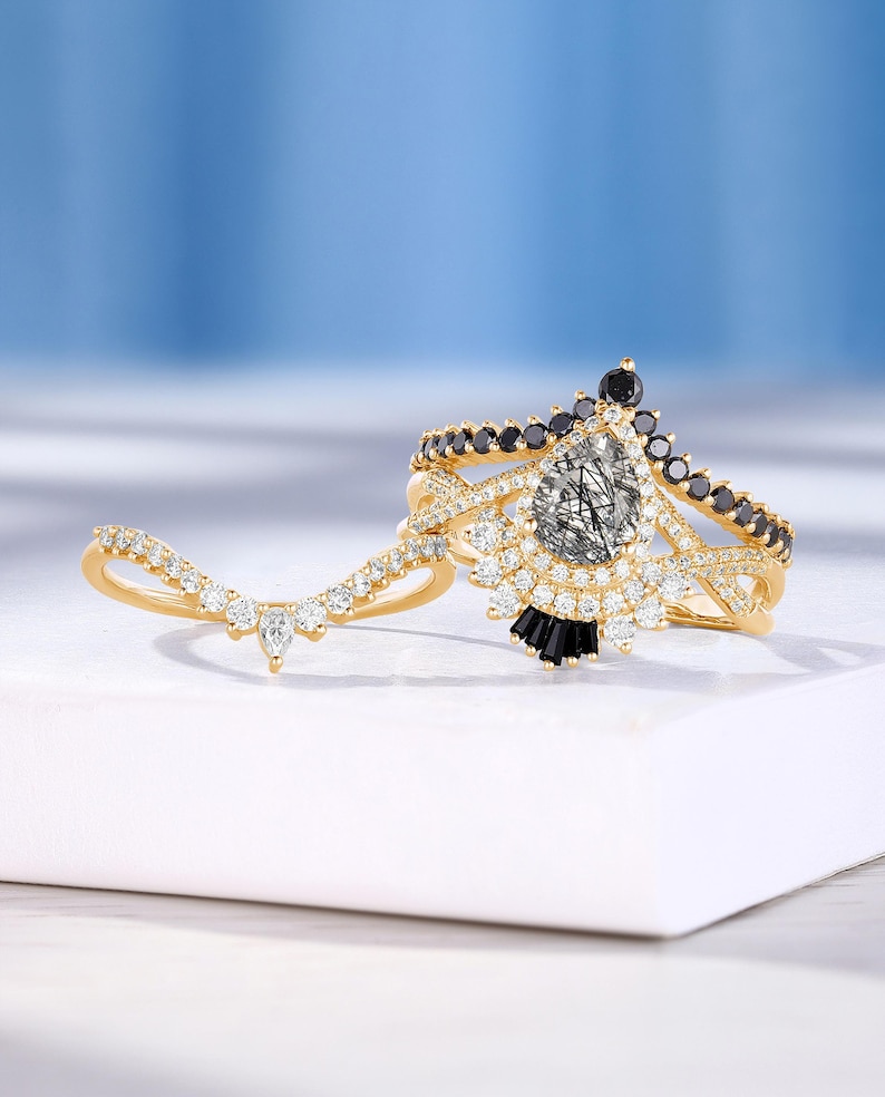 May include: A gold ring set with a pear-shaped clear gemstone surrounded by smaller clear gemstones and black gemstones. The ring is set on a white surface.