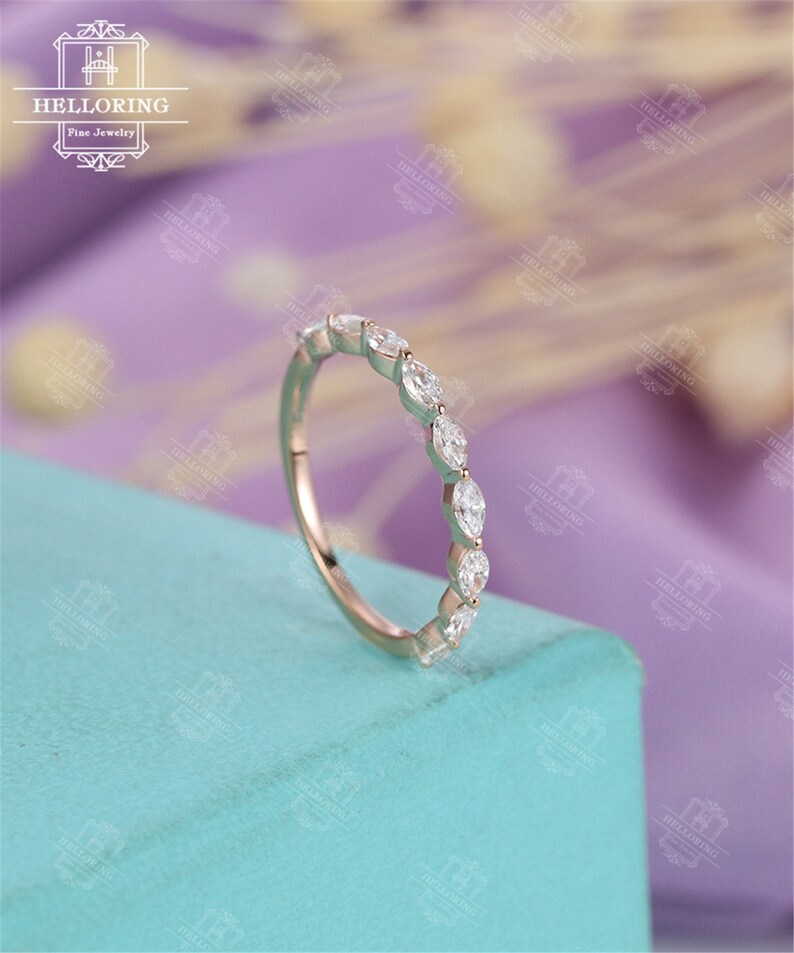 May include: A delicate, rose gold band with a row of marquise-shaped diamonds set in a prong setting. The band is on a light blue surface.