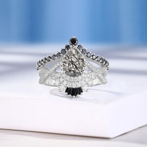 May include: A pear-shaped engagement ring with a clear, sparkly center stone surrounded by smaller clear stones and black stones. The ring has a delicate, intricate design with a crown-like setting.