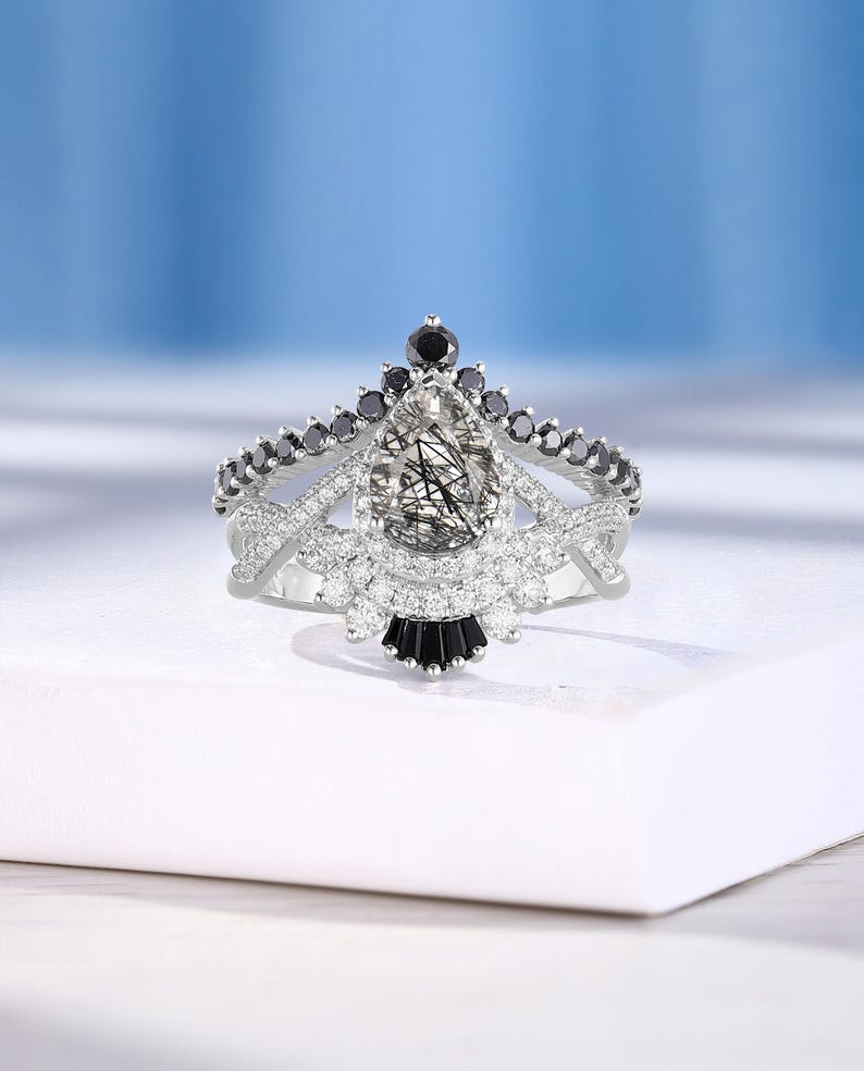 May include: A pear-shaped engagement ring with a clear, sparkly center stone surrounded by smaller clear stones and black stones. The ring has a delicate, intricate design with a crown-like setting.
