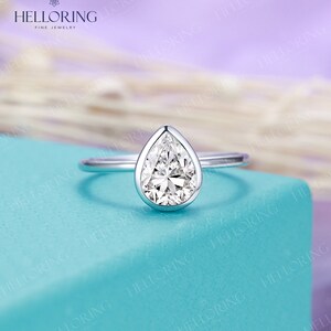 May include: A pear-shaped diamond engagement ring set in a white gold band. The ring is on a light blue background.