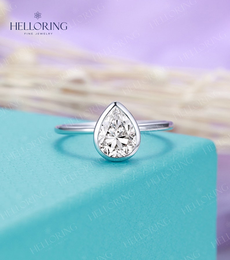 May include: A pear-shaped diamond engagement ring set in a white gold band. The ring is on a light blue background.