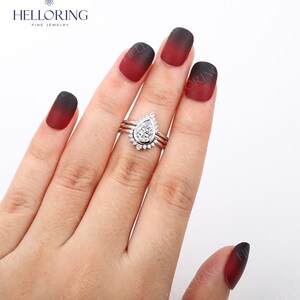 May include: A pear-shaped diamond engagement ring with a halo setting and two thin bands. The ring is on a hand with red and black ombre nail polish.