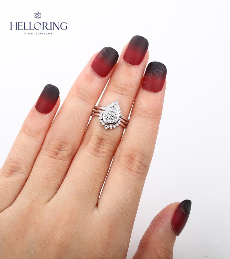 May include: A pear-shaped diamond engagement ring with a halo setting and two thin bands. The ring is on a hand with red and black ombre nail polish.