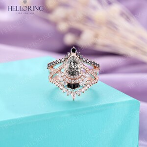 May include: A rose gold engagement ring with a pear-shaped grey and black diamond, surrounded by smaller diamonds. The ring has a delicate, intricate design with black diamonds and a three-band setting.