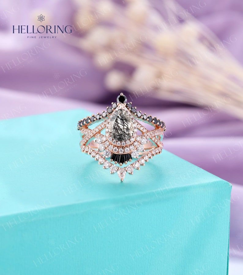 May include: A rose gold engagement ring with a pear-shaped grey and black diamond, surrounded by smaller diamonds. The ring has a delicate, intricate design with black diamonds and a three-band setting.