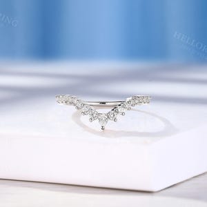 May include: A delicate, curved wedding band with a row of small, sparkling diamonds set in a white metal. The band is designed to fit snugly against a larger engagement ring.