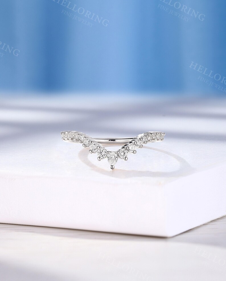 May include: A delicate, curved wedding band with a row of small, sparkling diamonds set in a white metal. The band is designed to fit snugly against a larger engagement ring.