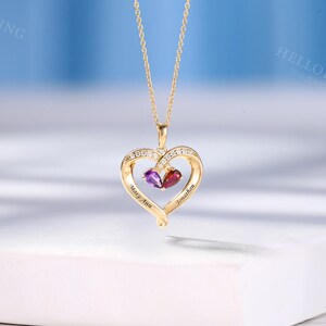 May include: A gold-coloured heart-shaped pendant necklace with a delicate chain. The heart is decorated with small clear stones and two gemstones, one purple and one red. The names "Mary Ann" and "Jonathan" are engraved on the heart.