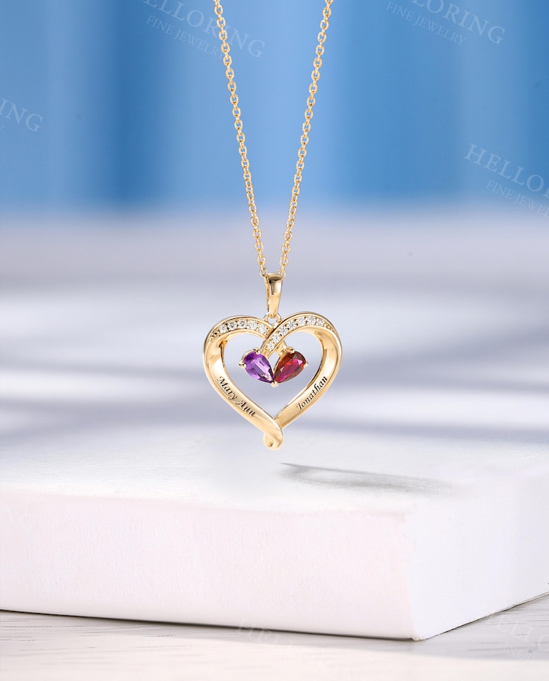 May include: A gold-coloured heart-shaped pendant necklace with a delicate chain. The heart is decorated with small clear stones and two gemstones, one purple and one red. The names "Mary Ann" and "Jonathan" are engraved on the heart.