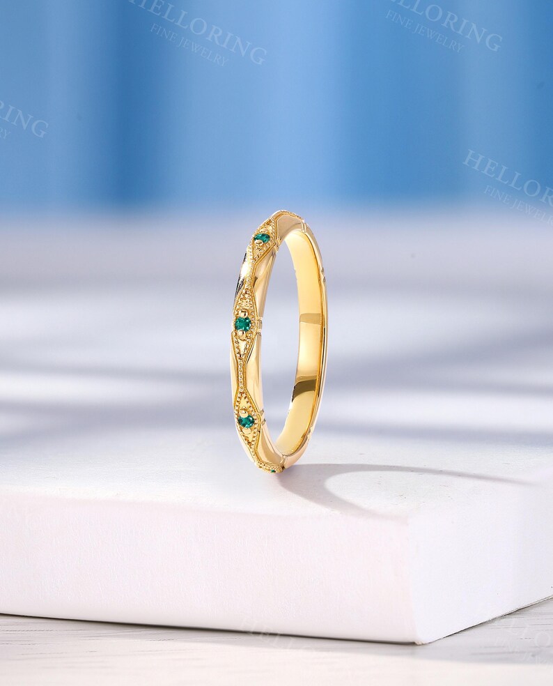 May include: A yellow gold band ring featuring a series of small, round, green gemstones set within decorative, diamond-shaped metal frames. The band has a polished finish and intricate milgrain detailing along the edges of the settings.