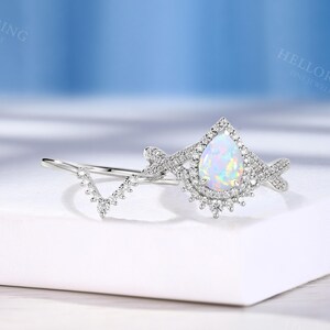 May include: A silver engagement ring set featuring a pear-shaped opal center stone surrounded by a halo of small, clear gemstones. The band is adorned with additional small gemstones. A matching V-shaped band is included in the set.