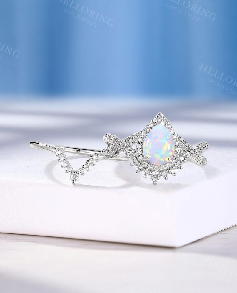 May include: A silver engagement ring set featuring a pear-shaped opal center stone surrounded by a halo of small, clear gemstones. The band is adorned with additional small gemstones. A matching V-shaped band is included in the set.