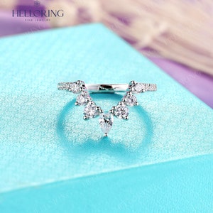 May include: A delicate silver ring with a V-shaped design featuring a row of small round diamonds and a larger pear-shaped diamond hanging from the center.