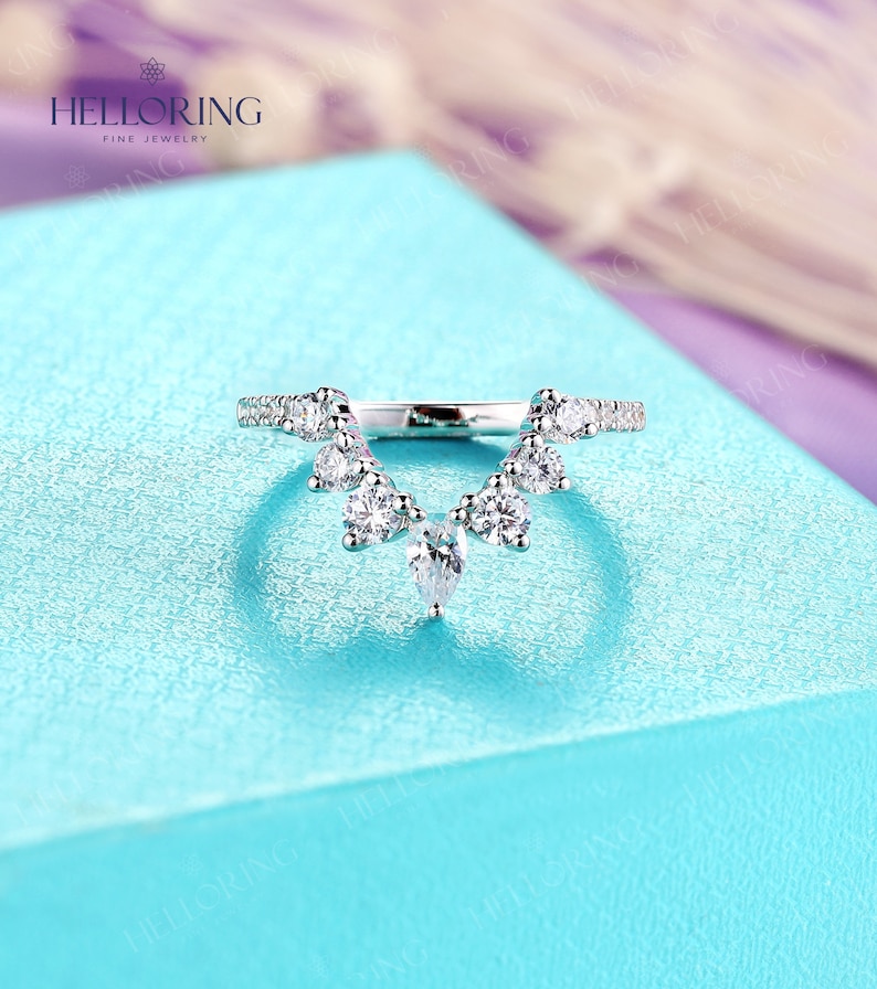 May include: A delicate silver ring with a V-shaped design featuring a row of small round diamonds and a larger pear-shaped diamond hanging from the center.