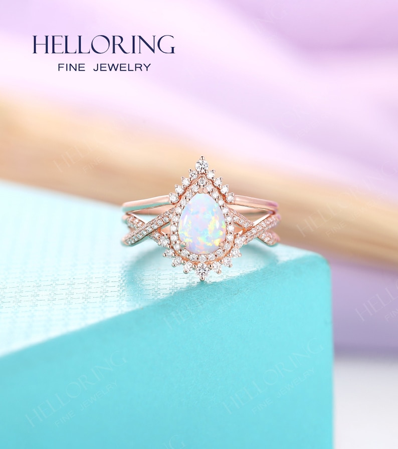 May include: A rose gold engagement ring with a pear-shaped opal gemstone surrounded by a halo of smaller diamonds. The ring is set on a delicate band with a criss-cross design.