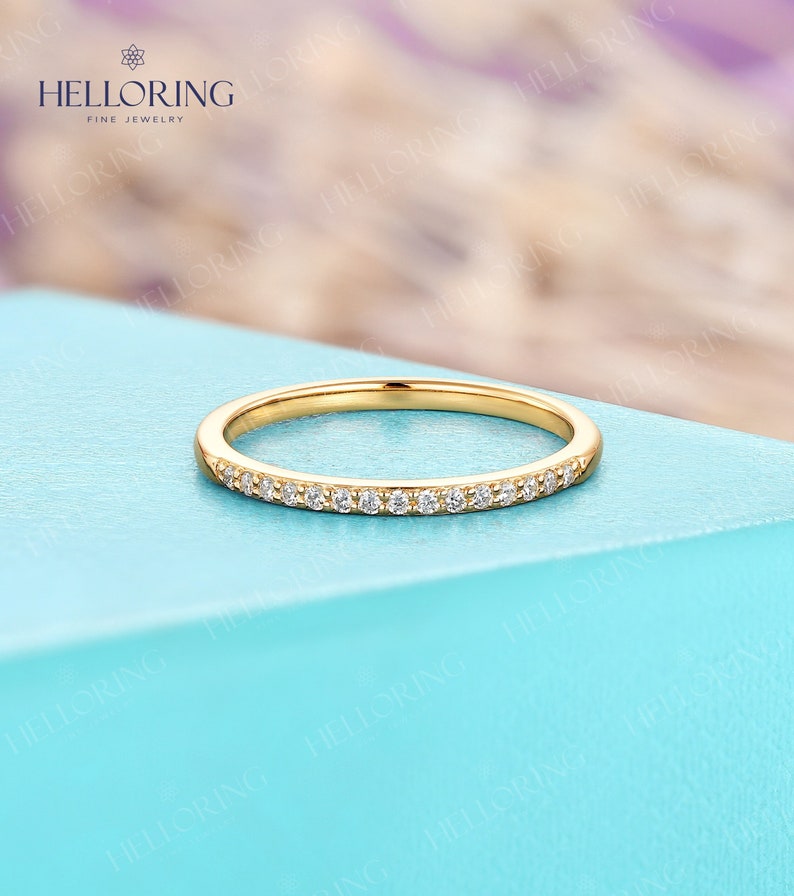 May include: A delicate gold band ring with small, clear gemstones set along the top. The ring is displayed on a light blue surface. The HELLORING FINE JEWELRY logo is visible in the background.